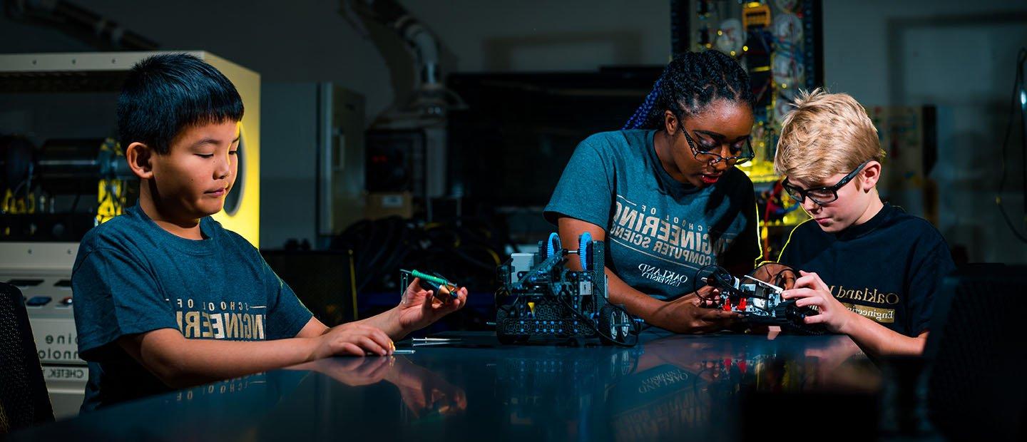 three kids standing around a table in a lab, working with mechanical equipment