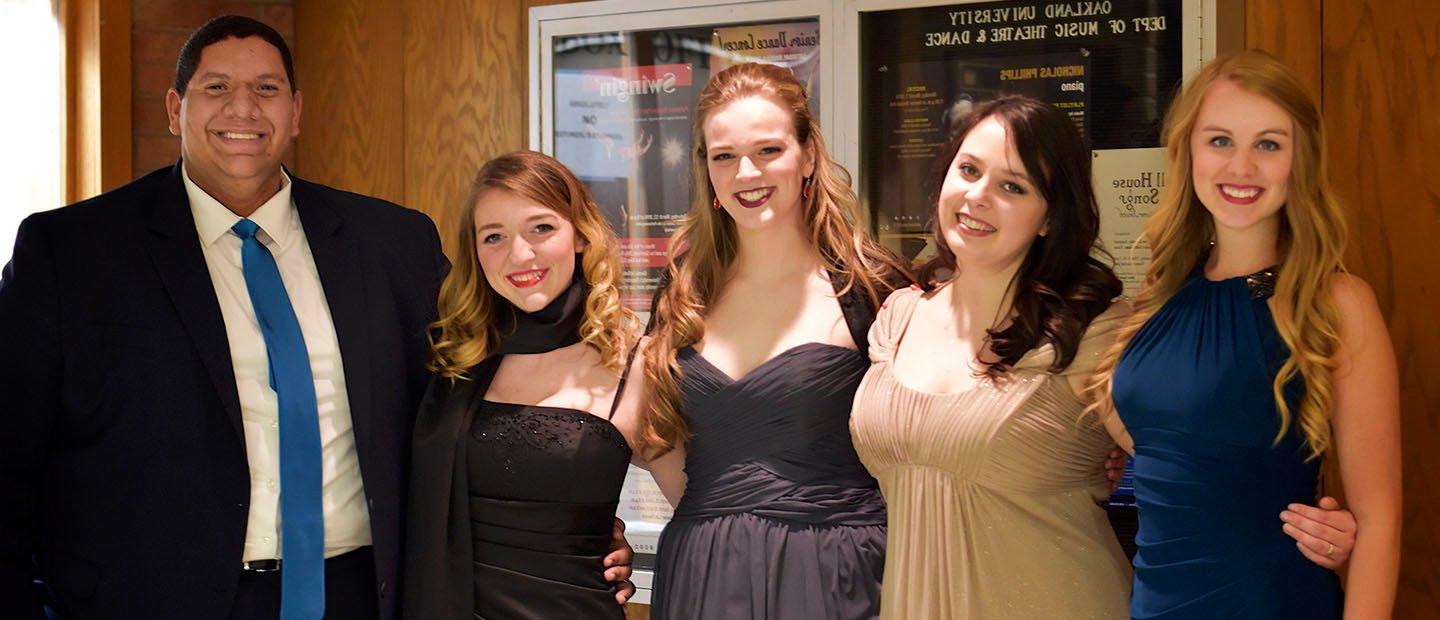 five people in evening attire, standing in a row with arms around each other, smiling at the camera