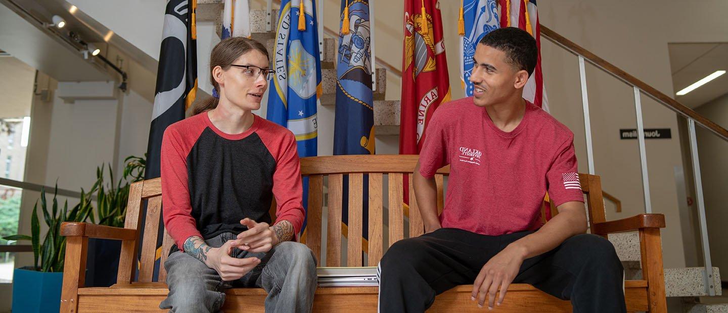 two people wearing red and black seated on a wooden bench with flags behind them